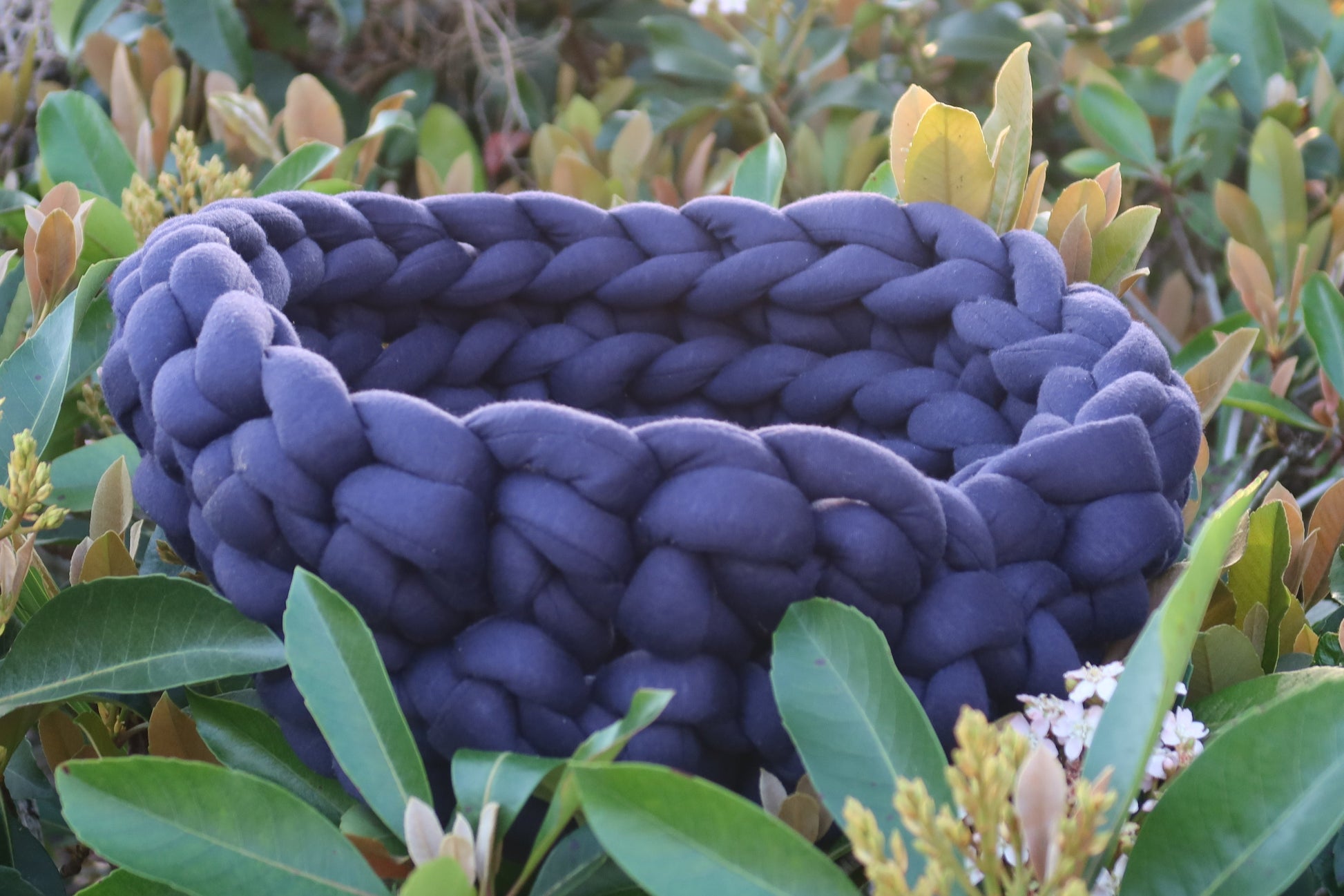 Navy blue braided dog bed in a natural setting with green foliage